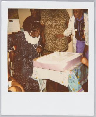 Elderly woman in patterned dress getting ready to blow out candles on cake]