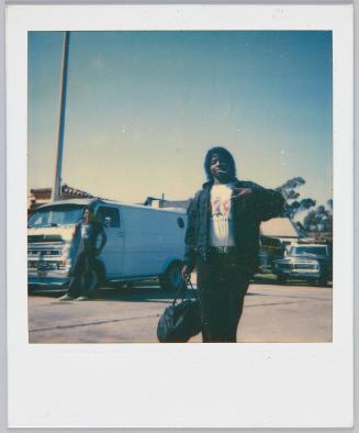 [Man with curly hair standing in parking lot holding up peace sign up and lollipop in mouth]