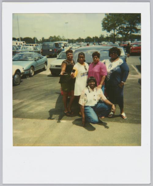 [Group of five woman posing in parking lot, two women on left holding polaroids]