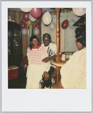[Woman in white and red patterned sweater sitting in between man's legs, red and white balloons suspended from the ceiling above them]