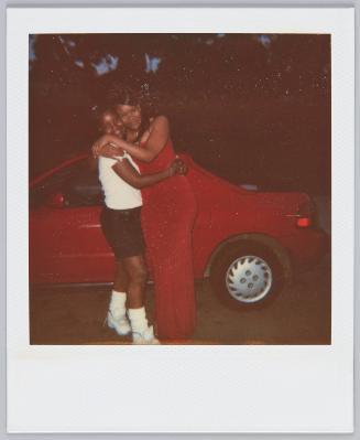 [Women in red outfit hugging younger girl, both standing in front of red car]