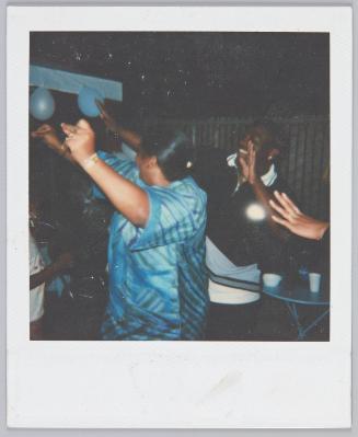 [People dancing with their arms in the air, woman in foreground wears silver blue top]