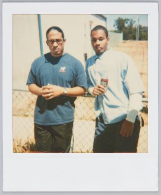 [Two young men leaning by metal fence, man on right holding a can of soda]