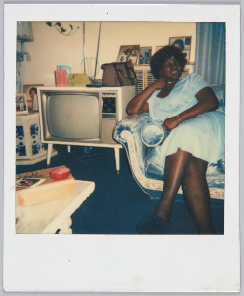 [Woman in blue dress sitting on couch leaning on hand and looking away from camera]