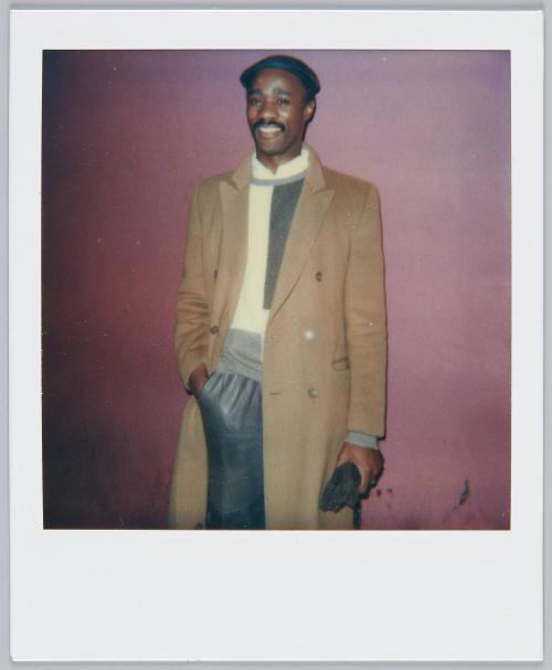 [Man with moustache wearing long brown trenchcoat standing in front of maroon wall]