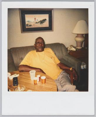 [Elderly man in yellow tshirt sitting on couch, painting of a lighthouse on the wall behind him]