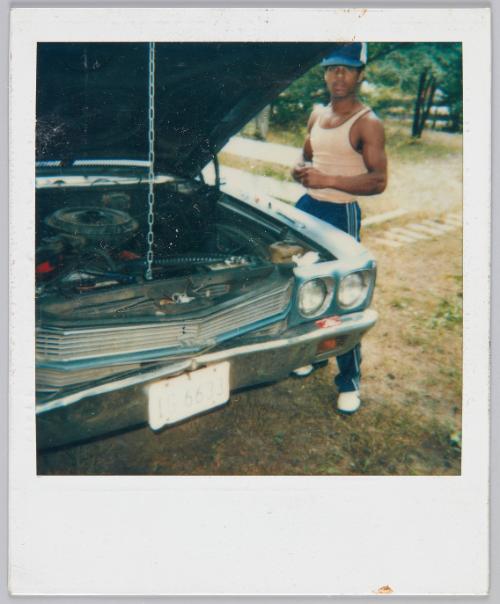 [Man in tank top and sweatpants standing next to car with the hood propped open]