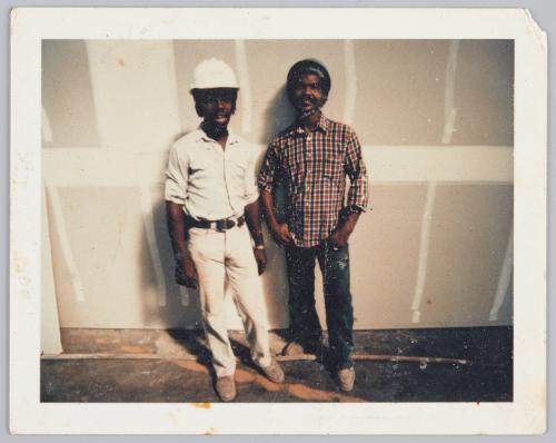 [Two men standing in front of wall under construction, man on left is wearing a white helmet]