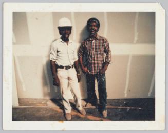 [Two men standing in front of wall under construction, man on left is wearing a white helmet]