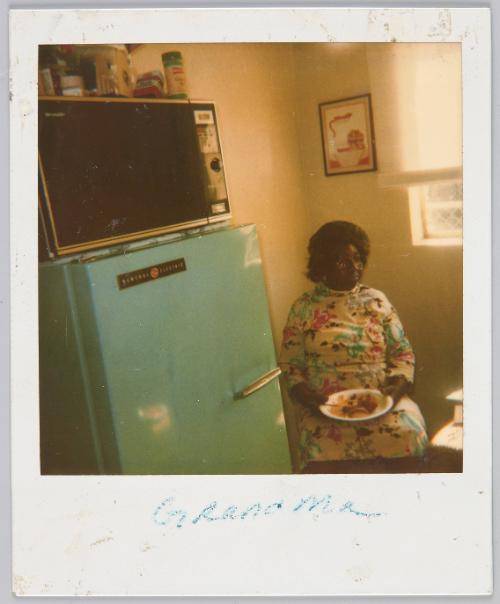 Grandma [Elderly woman sitting with plate of food on her lap next to green fridge]