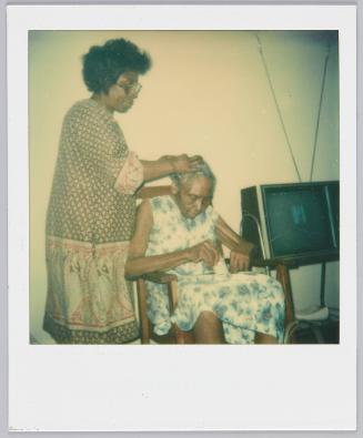 [Woman touching elderly woman's head while she sits in wooden chair beside television set]