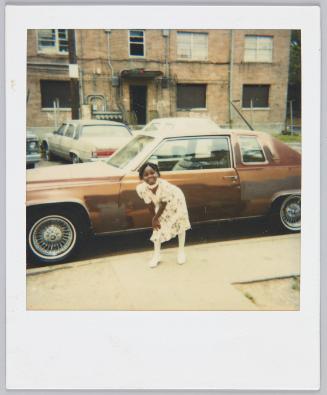 [Young girl in floral white dress standing on sidewalk beside brown car, leaning with both hands on her knee]