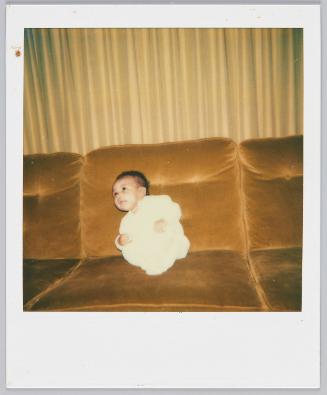 [Baby in white sitting on velvet couch looking up to the left]