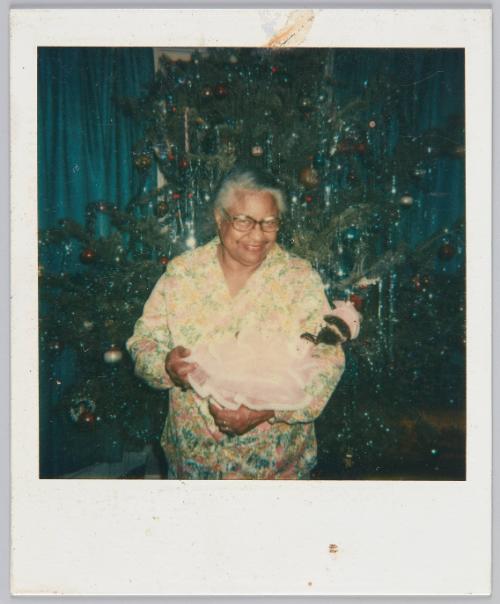 [Elderly woman smiling in her pajamas, standing in front of Christmas tree with doll in her arms]