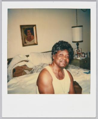 [Man in white tank top with silver necklace smiling and sitting on bed with white sheets, framed portrait above the bed]