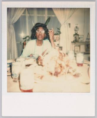 [Elderly woman wearing eyeglasses sitting at table with food and glasses on table, her left hand is raised]