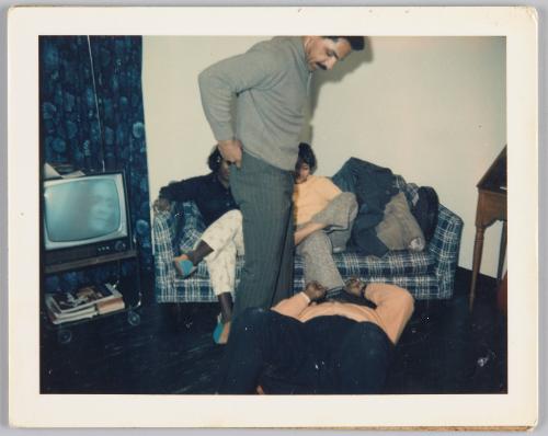 [Two women sitting on couch, man lying on floor in front of them with another man standing over him looking down]