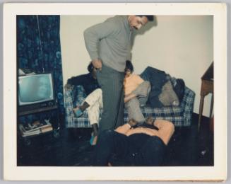 [Two women sitting on couch, man lying on floor in front of them with another man standing over him looking down]