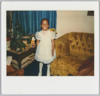 [Young girl in white formal dress, white stockings and Mary-Jane shoes standing and smiling in living room with golden velvet couch behind her]