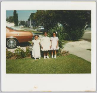 [Three young girls dressed in white standing in row on lawn in front of sidewalk, brown car in background]