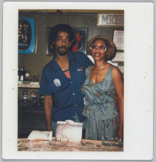 [Man and woman side by side standing behind bar with an advertisement on bartop 'White Turkey']

