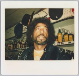 [Man standing in front of display case in store, black hats hung above him]
