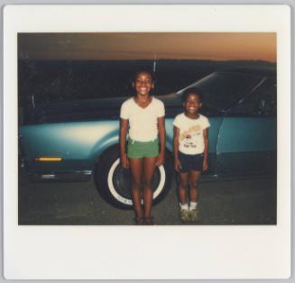 [Two girls standing side by side in front of car smiling with arms at their sides]
