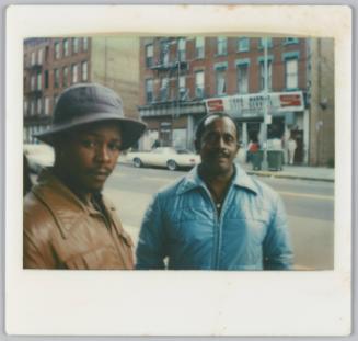 [Two men dressed in jackets standing on city street, food market in background]
