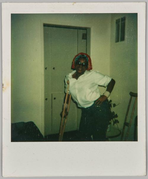 [Woman in white shirt leaning on crutch with her hand on her hip, standing in front of grey door]