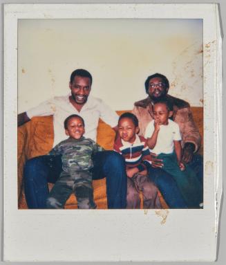 [Two men and three boys on orange couch, boy in centre in red, blue and white striped collared shirt and making funny face]