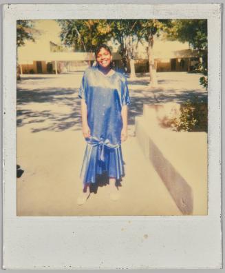 [Woman in blue dress and white shoes smiling and standing outside by cement planters]