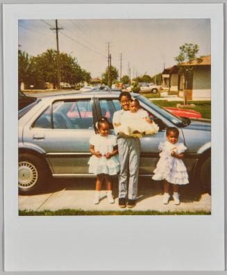 [Boy in grey dress pants holding baby in white dress, two young girls in white dresses standing beside him in driveway]