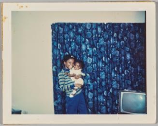[Young woman in striped shirt holding a baby and standing in front of a blue floral curtain]