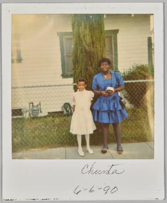 Chienta 6-6-90 [Girl in white dress standing beside a woman in a blue dress, outside in front of fence and tree trunk]