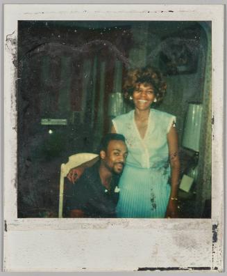 [Woman in blue dress standing with her arm around a man sitting down]