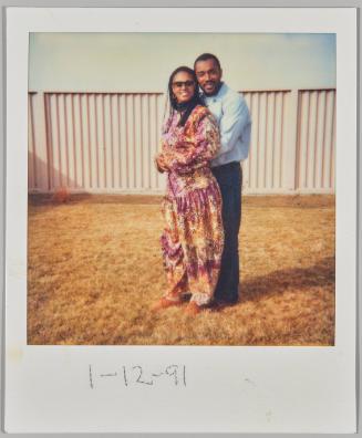 1-12-91 [Man in blue shirt holding woman in sunglasses on a lawn with a tall white fence]