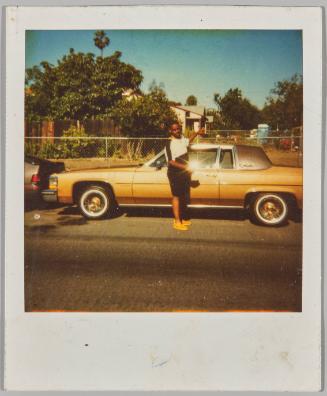 [Man standing by gold car parked on the road in front of chainlink fence in residential neighbourhood]