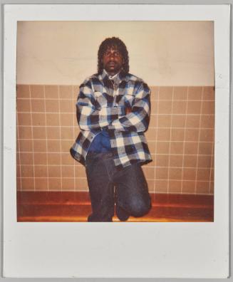 [Man in blue, white and black plaid shirt leaning against white wall with beige tiles]