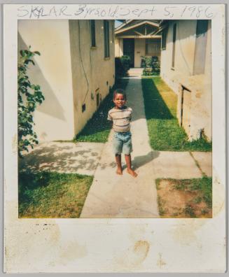 Skylar 3 yrs. old Sept 5, 1986 [Boy standing barefoot on walkway between houses]