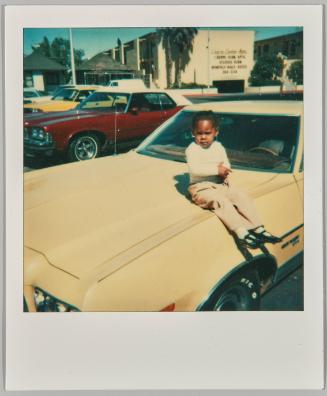 [Child in a white holding their hands together and sitting on the hood of car in parking lot]