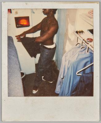 [Profile of shirtless man with cigarette in mouth, to the right are shirts on hangers]