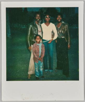 [Three adults and a child standing together, woman in centre in sunglasses and white shirt]