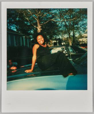 [Woman dressed in black sitting on hood of car, trees in background]