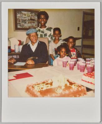 [Elderly man in cap and glasses sitting, woman and children standing beside him and a table with cake]