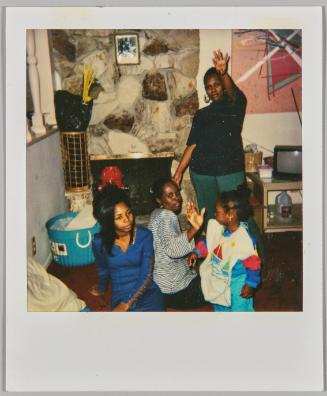 [Two women sitting on floor in front of stone fireplace, waving woman and girl standing to the right]