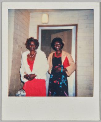 [Woman in red dress and white coat with woman in patterned dress and beige coat, standing outside door]