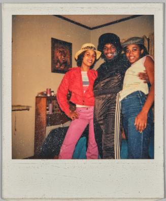 [Two adolescent women and adolescent man standing together in home, all wearing hats, woman on left wearing a red jacket and pink pants]