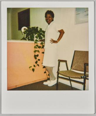 [Woman in white shirt and dress, wearing tennis shoes and smiling with hand on hip next to leafy plant]