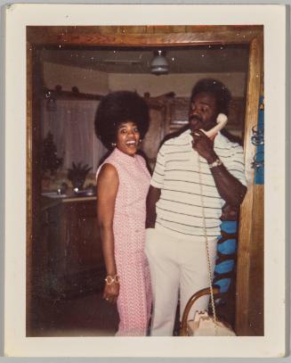 [Woman in pink jumpsuit smiling with eyes wide next to man talking on telephone]