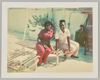 [Woman in red jumpsuit and another woman in pink pants sitting together outside on a sunchair, potted plant and wall with outlet behind them]